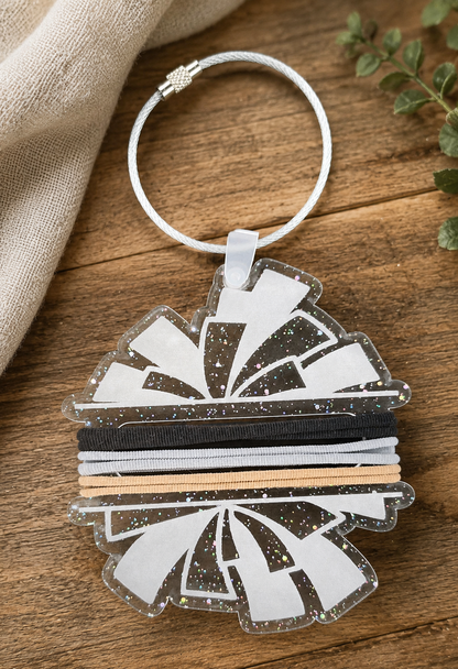 A decorative acrylic keychain featuring pom shaped design with alternating white and glittery segments, attached to a wire ring , displayed on a rustic wooden surface with a linen cloth and green leaves.