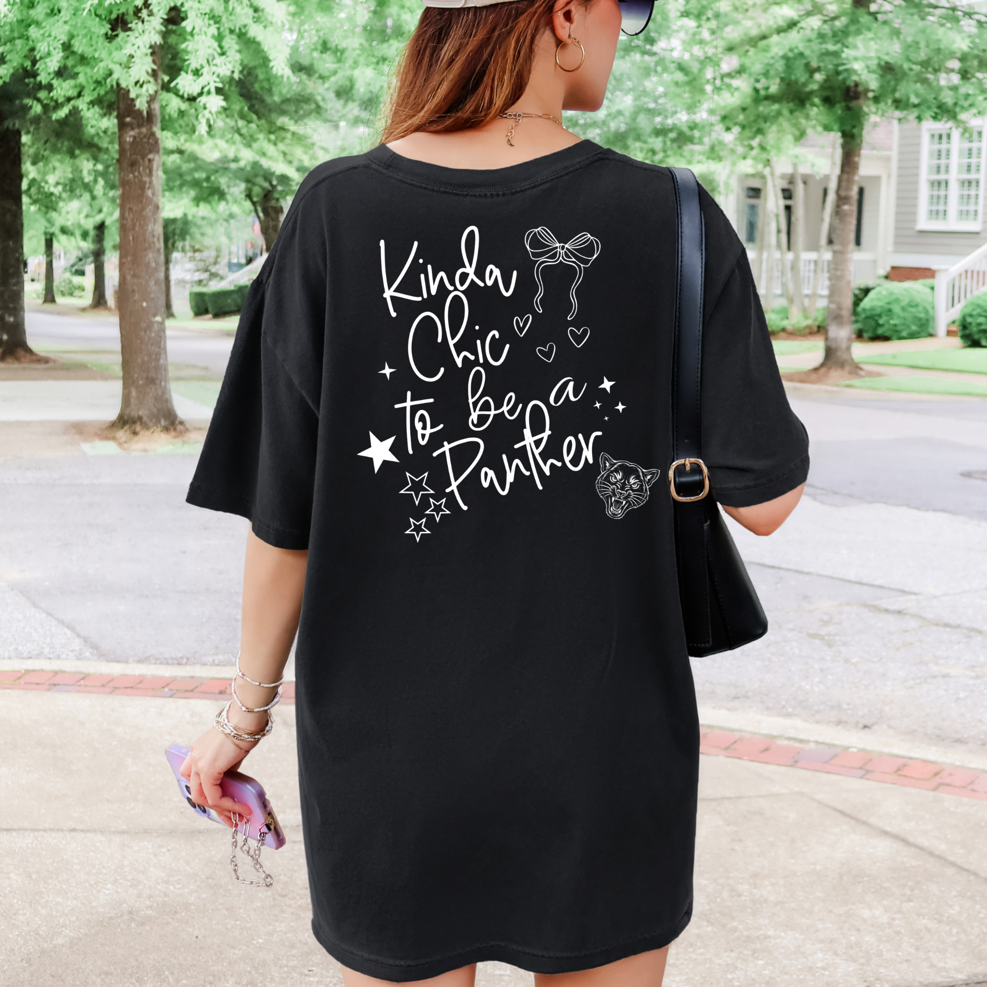 A woman wearing a black oversized T-shirt with white graphic text that reads 'Kinda Chic to be a Panther' and small white decorative illustrations, photographed outdoors on a sidewalk with trees and houses in the background.