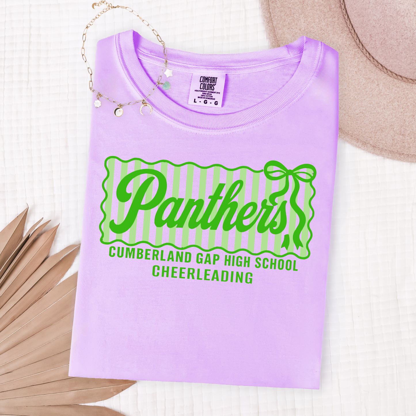 A folded light purple t-shirt with a green and white striped 'Panthers' graphic, displayed on a white textured background with a straw hat, necklace, and dried palm leaves.