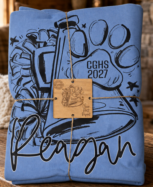 A folded light blue blanket with a black graphic print of a paw print, the text 'CGHS 2027', and the name 'Reagan' in script. The shirt is wrapped in twine with a wooden tag.